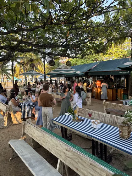 Mesa longa montada para um evento na areia, com toalhas listradas